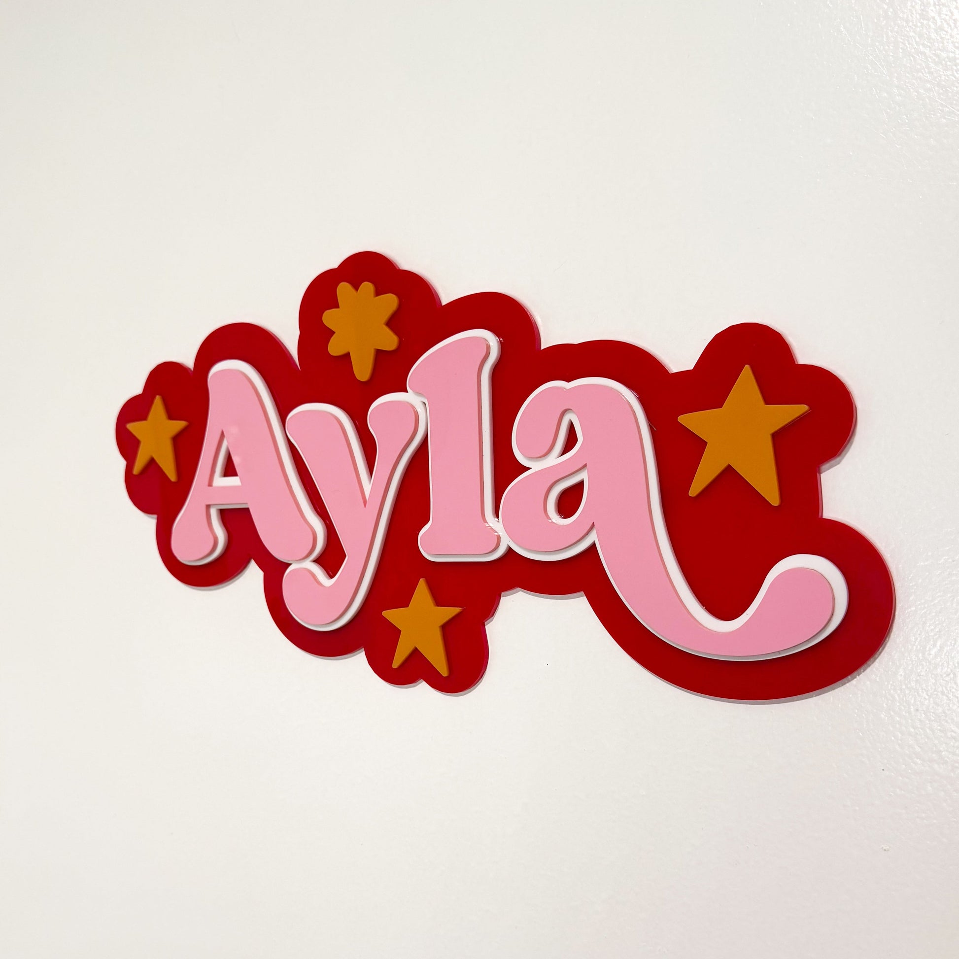 Decorative nameplate with 'Ayla' in pink on a red background with yellow stars, on a white background.